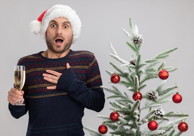 Geïmponeerde jonge blanke man met kerstmuts staande in de buurt van de kerstboom met glas champagne kijken camera hand houden op de borst geïsoleerd op een witte achtergrond