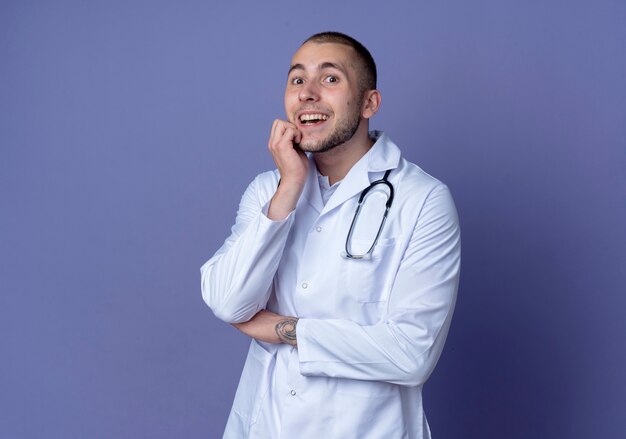 Geïmponeerde jonge mannelijke arts die medische gewaad en stethoscoop draagt die handen op kin en onder elleboog zetten die op paars wordt geïsoleerd