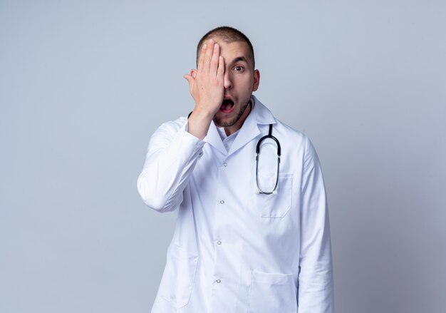 Geïmponeerde jonge mannelijke arts die medische mantel en stethoscoop om zijn nek draagt die de helft van het gezicht bedekt met hand die op wit wordt geïsoleerd