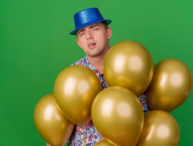 Gratis foto geïmponeerde jonge partijkerel die blauwe hoed draagt die zich onder ballons bevindt die op groen worden geïsoleerd