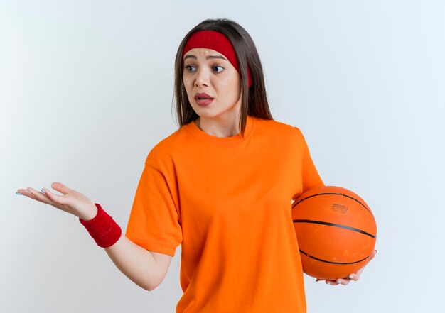 Geïmponeerde jonge sportieve vrouw die hoofdband en polsbandjes draagt die lege hand tonen die de bal van het zijholdingbasketbal bekijken