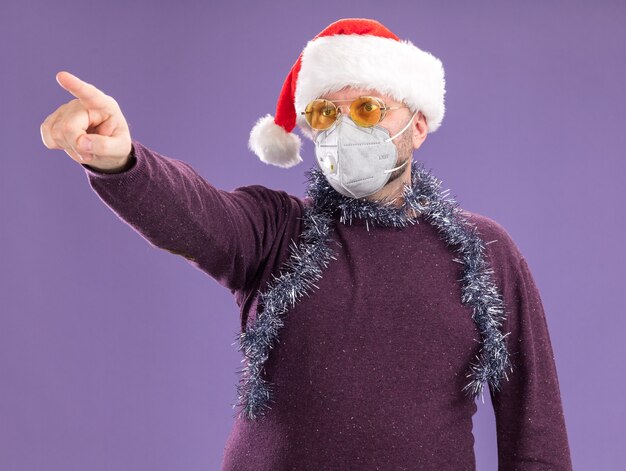 Geïmponeerde man van middelbare leeftijd met kerstmuts en beschermend masker met klatergoud slinger om nek met bril kijken en wijzend naar kant geïsoleerd op paarse muur