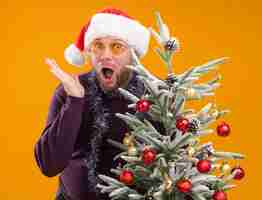 Gratis foto geïmponeerde man van middelbare leeftijd met kerstmuts en klatergoud slinger om de nek met een bril achter versierde kerstboom met lege hand geïsoleerd op een oranje muur