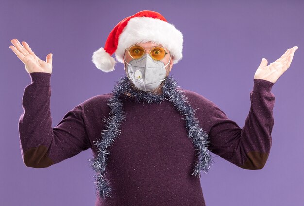 Geïmponeerde man van middelbare leeftijd met kerstmuts en klatergoud slinger rond de nek met bril en beschermend masker camera kijken