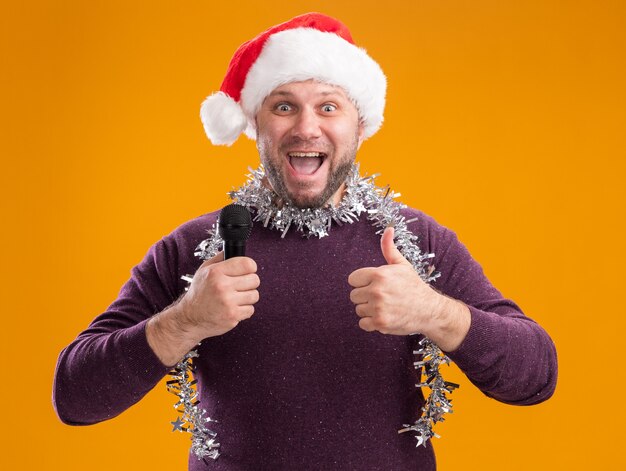 Geïmponeerde man van middelbare leeftijd met kerstmuts en klatergoud slinger rond de nek met microfoon die duim toont die omhoog op oranje muur wordt geïsoleerd
