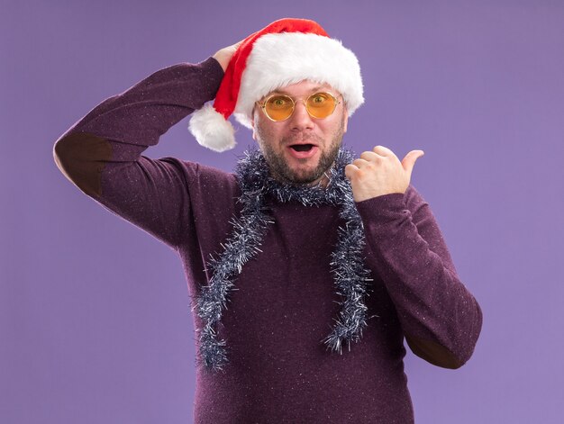 Geïmponeerde man van middelbare leeftijd met kerstmuts en klatergoudslinger om de nek met een bril die de hand op het hoofd houdt en naar de zijkant wijst geïsoleerd op paarse muur