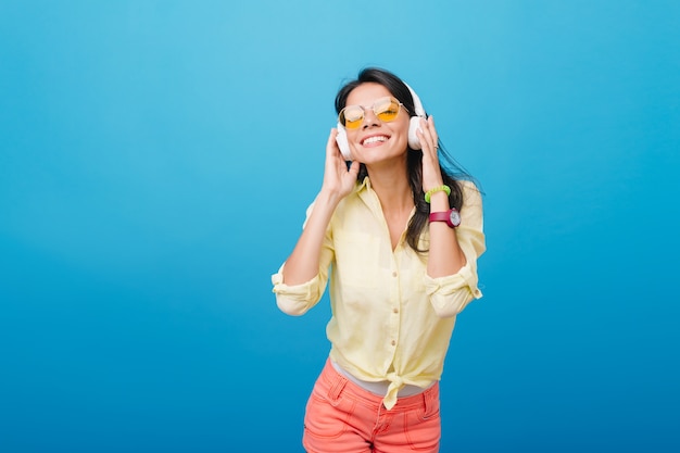 Geïnspireerd Aziatisch vrouwelijk model in roze polshorloge en groene armband luisteren muziek. Indoor foto van extatisch Latijns meisje in oranje zonnebril koptelefoon aan te raken en glimlachen.