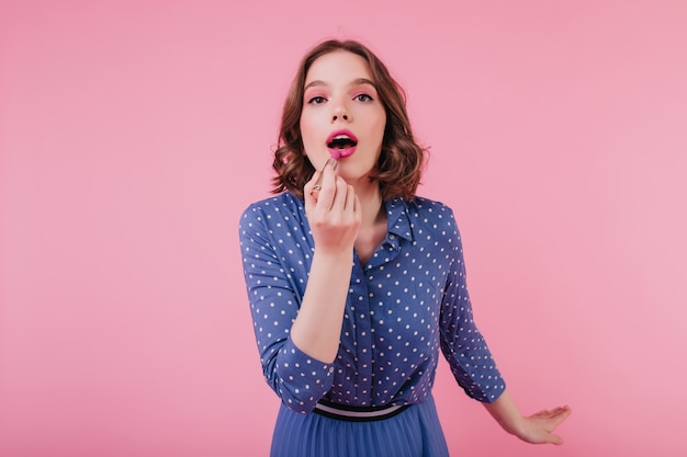 Geïnspireerd brunette meisje met donkere ogen met roze lippenstift. Indoor foto van modieuze jonge dame met kort kapsel doet haar make-up.