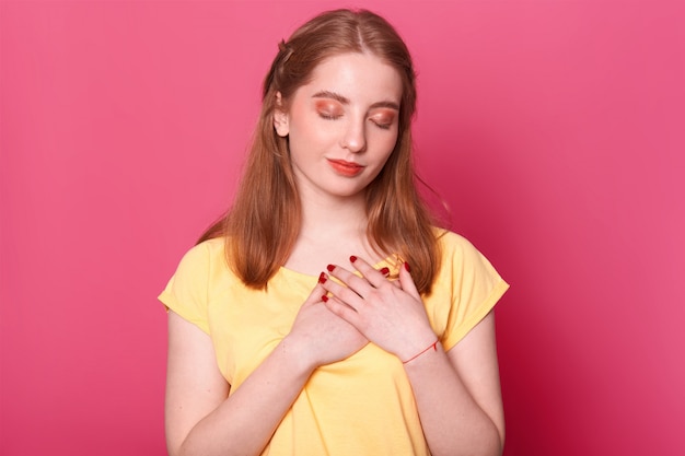 geïnspireerd kalm roodharig wit meisje met rode lippenstift op haar lippen, manicure, make-up en mooi kapsel staat met gekruiste handen op haar hart met gesloten ogen, lijkt attent te zijn.