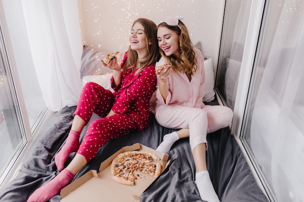 Geïnspireerd meisje in roze sokken pizza eten met beste vriend. Binnenfoto van twee zusters in pyjama die van Italiaans eten in bed genieten.