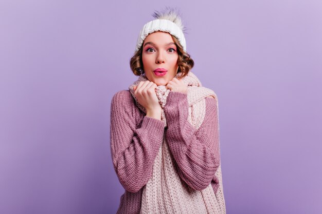 Geïnspireerd meisje met roze make-up en verraste glimlach die zich voordeed op paarse muur. schattige jonge vrouw in hoed en sjaal geluk uitdrukken.