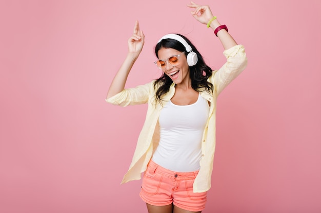 Geïnspireerd slank brunette meisje in zonnebril grappige dansende en zwaaiende handen. Lachen donkerharige jonge vrouw in geel overhemd genieten van muziek in koptelefoon met gesloten ogen.