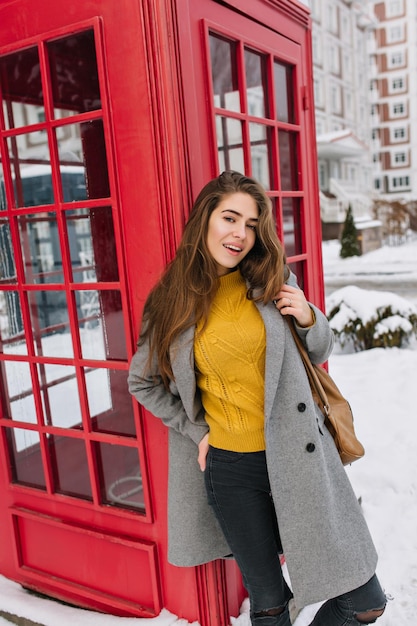 Geïnspireerd vrouwelijk model in grijze jas die in decemberdag naast de telefooncel staat met een zachte glimlach. Outdoor portret van prachtige Europese vrouw in gele trui genieten van winterochtend op straat.