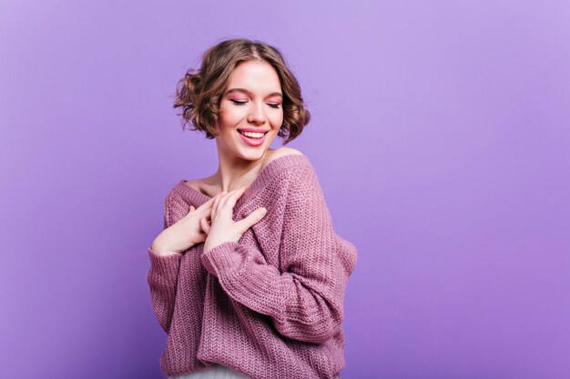 Geïnspireerde blanke vrouw draagt een zachte gebreide trui poseren met een verlegen glimlach. Indoor portret van enthousiaste jonge dame met trendy kapsel lachen op paarse muur.