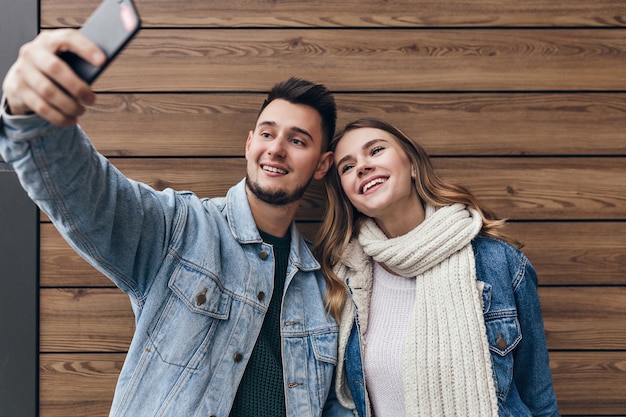 Geïnspireerde man met baard selfie maken met zijn vriendin. Prachtige jonge vrouw met zwarte sjaal poseren op houten muur.