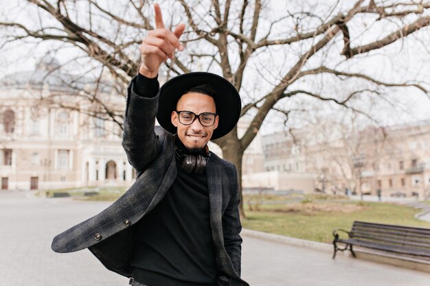 Geïnspireerde zwarte man in grijze jas hand zwaaien in park. Openluchtportret van blij Afrikaans mannelijk model in hoed en glazen die op stadsplein rusten.