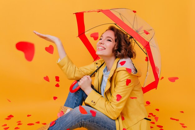 Geïnteresseerd bruinharig meisje dat van hartregen geniet. Glimlachende aantrekkelijke vrouw in gele jas zittend op de vloer met parasol.