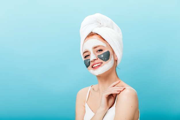Geïnteresseerd meisje met gezichtsmasker camera kijken. Studio die van goedgehumeurde dame met handdoek op hoofd is ontsproten die huidverzorgingsbehandeling doet.