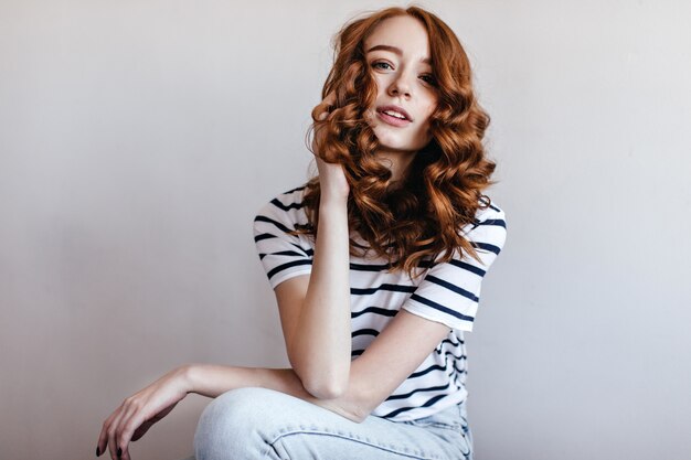 Geïnteresseerde gembervrouw in en jeans zitten kijken. Indoor foto van goedgehumeurd roodharig meisje in trendy casual t-shirt.