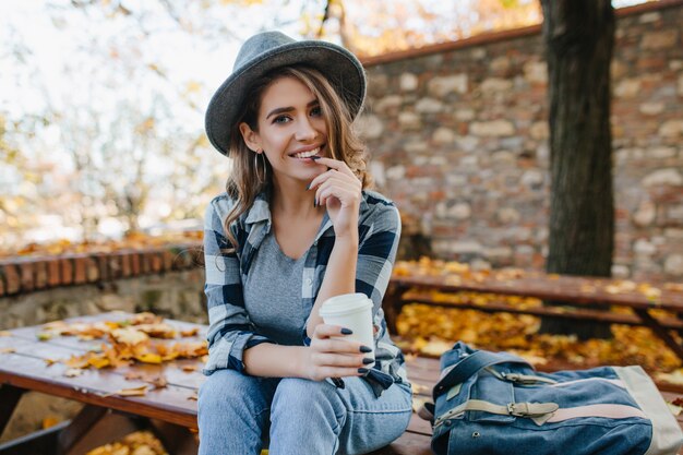 Geïnteresseerde jonge dame met kopje koffie poseren in park in oktober dag