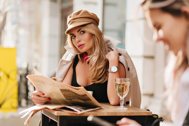 Geïnteresseerde jonge vrouw rondkijken, krant vasthouden en wijn drinken. Outdoor Portret van mooi meisje draagt GLB en stijlvolle beige jas in koude dag tijdens rust in café.