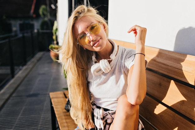 Geïnteresseerde langharige vrouw die zich voordeed op houten bankje in zonnige dag. Buiten schot van mooie blonde vrouw in grote koptelefoon.