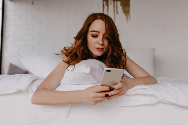 Geïnteresseerde roodharige vrouw die in bed ligt en naar het telefoonscherm kijkt. Lui vrouwelijk model met gemberhaar poseren in slaapkamer.