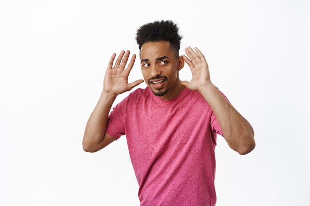 Geïntrigeerde Afro-Amerikaanse man die roddels probeert te horen, handen in de buurt van oren houdt en zijwaarts kijkt, probeert iets op een luide plaats te horen, staande in roze t-shirt op wit