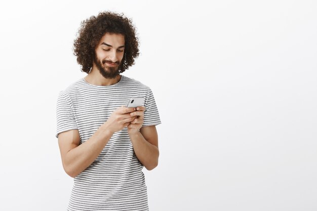 geïntrigeerde vrolijke hete Spaanse man met baard en afro kapsel, glimlachend op scherm van smartphone