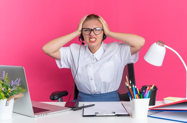 Geïrriteerd jong blond callcentermeisje met hoofdtelefoon en bril zittend aan een bureau met uitrustingsstukken die de handen op het hoofd houden en kijken naar de camera met tanden die op roze muur worden geïsoleerd