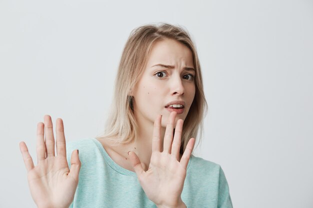 Geïrriteerde, bang, blonde, jonge vrouw met recht blond haar in blauwe trui die tegen de muur poseert, de handen in een stopgebaar houdt, zichzelf probeert te verdedigen en zegt: Stop daarmee. Lichaamstaal.