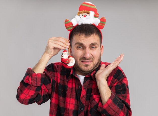 Geïrriteerde jonge blanke man met hoofdband van de kerstman kijken camera bedrijf kerstman kerst ornament in de buurt van hoofd met lege hand geïsoleerd op witte achtergrond