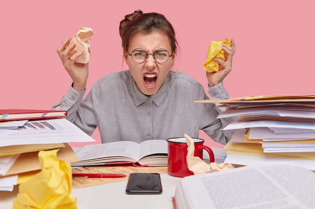 Geïrriteerde jonge blanke werknemer houdt papier bij, opent mond wijd, fronst gezicht, draagt shirt en bril, leest leerboek, analyseert documentatie