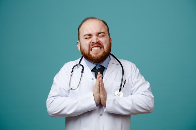 Geïrriteerde jonge mannelijke arts met medische jas en stethoscoop om zijn nek met namaste gebaar met gesloten ogen geïsoleerd op blauwe achtergrond