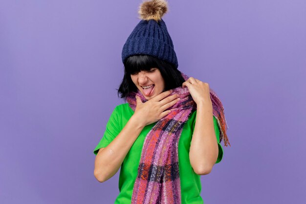 Geïrriteerde jonge zieke vrouw die de winterhoed en sjaal draagt die keel houdt neerkijkt geïsoleerd op purpere muur met exemplaarruimte
