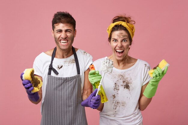 Geïrriteerde man en vrouw van schoonmaakdienst die vuile kleren dragen met reinigingsapparatuur die hun wenkbrauwen fronsen, bezig zijn met schoonmaken, kijken naar slordige meubels met walging en woede