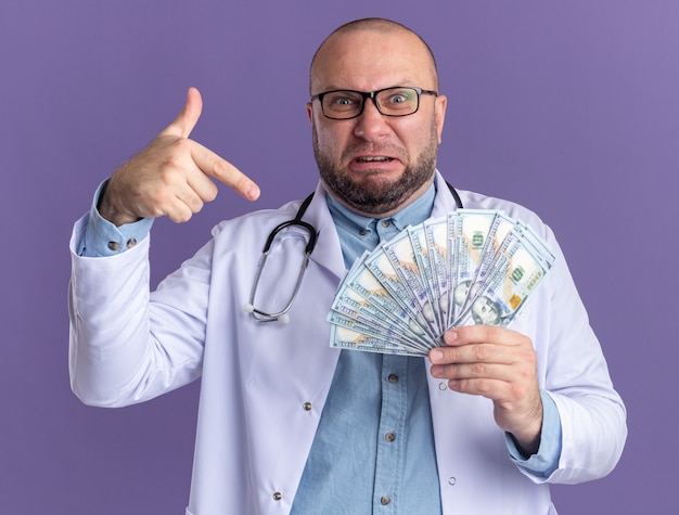 Geïrriteerde mannelijke arts van middelbare leeftijd die medische mantel en stethoscoop draagt met een bril die vasthoudt en wijst naar geld geïsoleerd op paarse muur