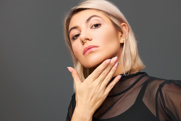 Geïsoleerd beeld van aantrekkelijke modieuze jonge vrouwelijke model met gezicht piercing en stijlvolle make-up poseren dragen trendy zwarte transparante blouse. Vrouwenstijl, mode en glamourconcept