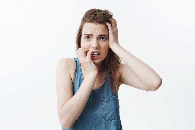 Geïsoleerd portret van mooi ongelukkig jong Kaukasisch studentenmeisje dat met donker lang haar knagende handen, voorhoofd houdt, dat opzij met schrikken uitdrukking kijkt.