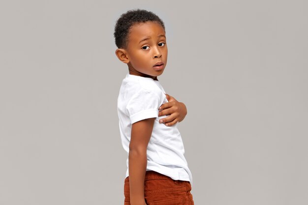 Geïsoleerd portret van perplex donkere jongen gekleed in wit t-shirt. Knap Afrikaans kind poseren. Lichaamstaal