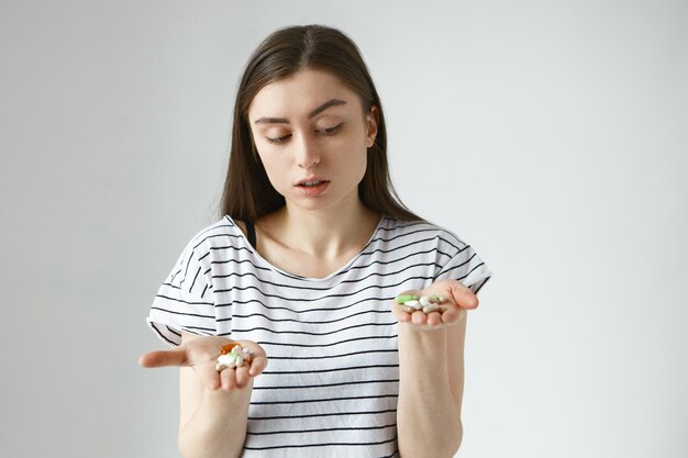 Geïsoleerd portret van twijfelachtige peinzende jonge Europese vrouw poseren met open handen voor haar, met een heleboel veelkleurige pillen op haar handpalmen, kiezen tussen antibiotica en vitamines