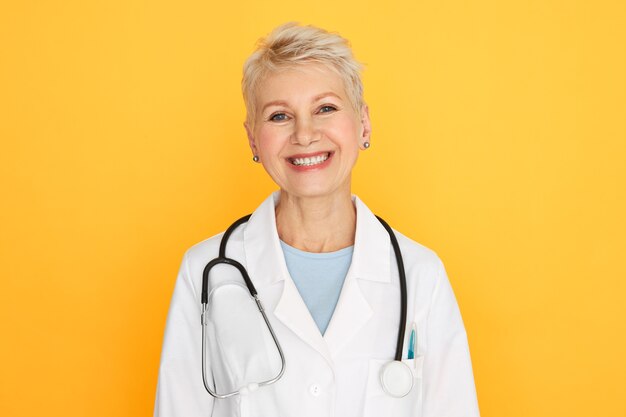 Geïsoleerd portret van zelfverzekerde ervaren vrouwelijke arts van middelbare leeftijd met kort blond kapsel die met gelukkige glimlach kijken