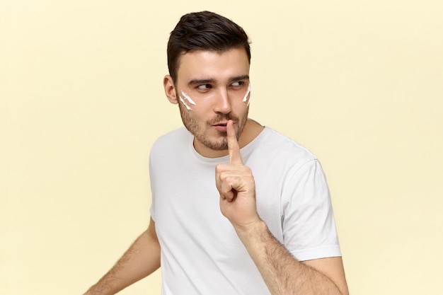 Geïsoleerde afbeelding van mysterieuze jonge man gekleed in een casual wit t-shirt met wijsvinger op de lippen, vragen om stil te zijn en zijn geheim niet aan andere mensen te vertellen.