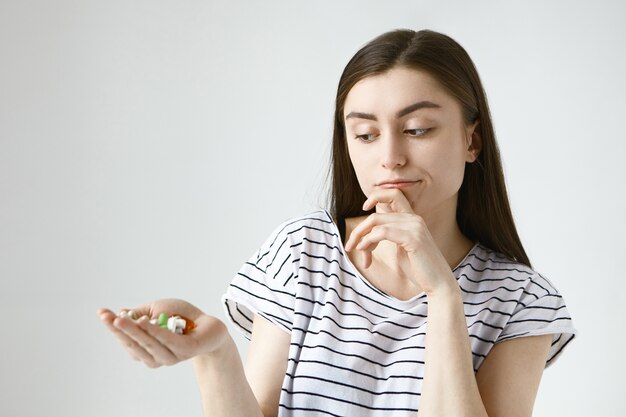 Geïsoleerde besluiteloze twijfelachtige jonge dame met losse donkere haren, bedachtzaam over de kin wrijven en kleurrijke pillen op haar handpalm bekijken, ze gaan nemen