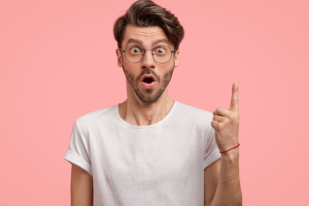 Geïsoleerde schot van verbaasde ongeschoren man hapt van verbazing, wijst met wijsvinger naar boven, draagt een ronde bril en wit t-shirt, modellen tegen roze muur, toont iets hierboven