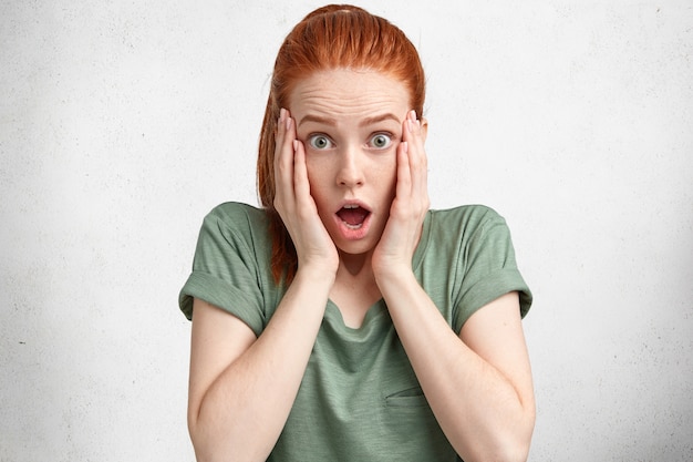 Geïsoleerde shot van bange emotionele vrouw met rood haar, houdt de handen op de wangen, voelt zich geschokt als iets vergeet, gekleed in een casual wit t-shirt, geïsoleerd over wit