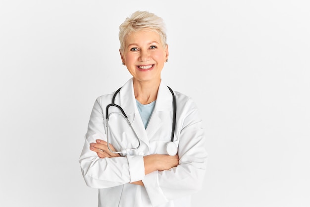 Geïsoleerde shot van een gelukkige succesvolle volwassen senior arts die medische unifrom draagt en een stethoscoop met vrolijke gelaatsuitdrukking, breed glimlachend, de armen gekruist op de borst