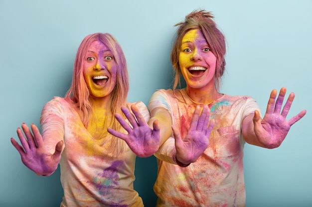 Geïsoleerde shot van emotionele gelukkige vrouwen strekken hun handen uit en tonen gekleurde handpalmen, lachen en hebben plezier binnen, vieren festival van kleuren, staan tegen een blauwe muur. holi-feest en feest