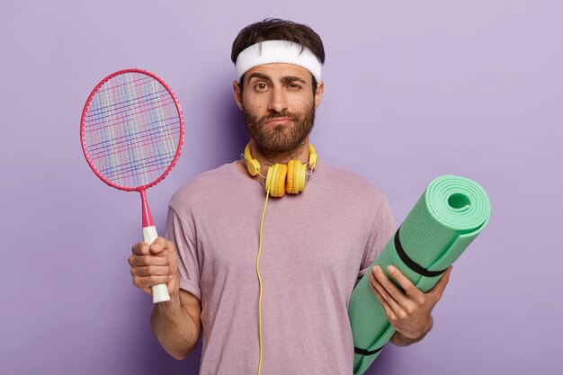 Geïsoleerde shot van ernstige man klaar voor het spelen van tennis tijdens tijdverdrijf, houdt racket en karemat, luistert naar muziek