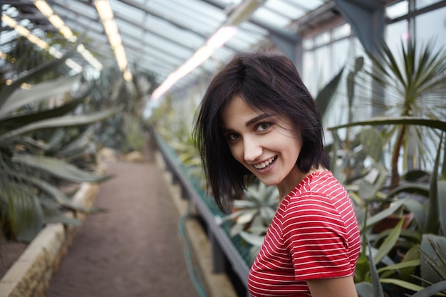 Geïsoleerde shot van mooie charismatische jonge brunette vrouw van in de twintig die vrolijk lacht naar de camera tijdens het wandelen in de plantenkwekerij tussen verschillende tropische bomen en struiken, zich zorgeloos voelt